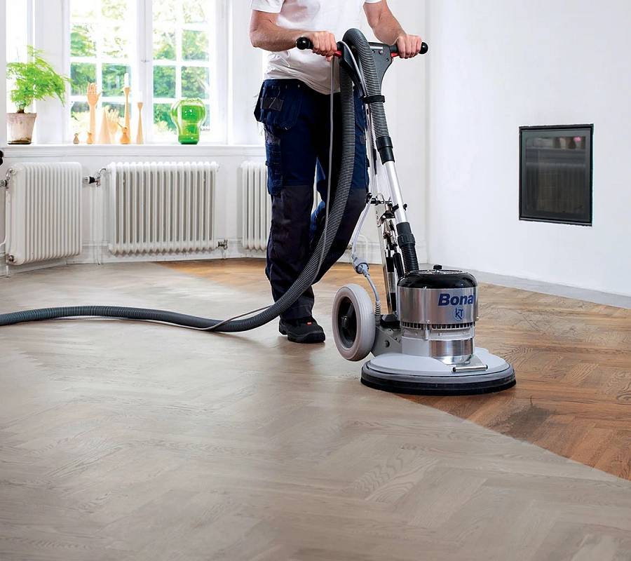 Handwerker schleift hellen Parkettboden mit Bodenschleifmaschine in leerem Raum