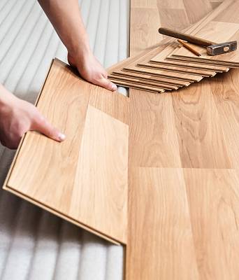 Hände verlegen Laminat im Fischgrätmuster passgenau, daneben liegen Hammer und Zubehör für die fachgerechte Montage.