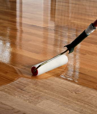 Auftragen einer schützenden Versiegelung mit Farbrolle auf einen Holzparkettboden, glänzende Oberfläche während der Behandlung.