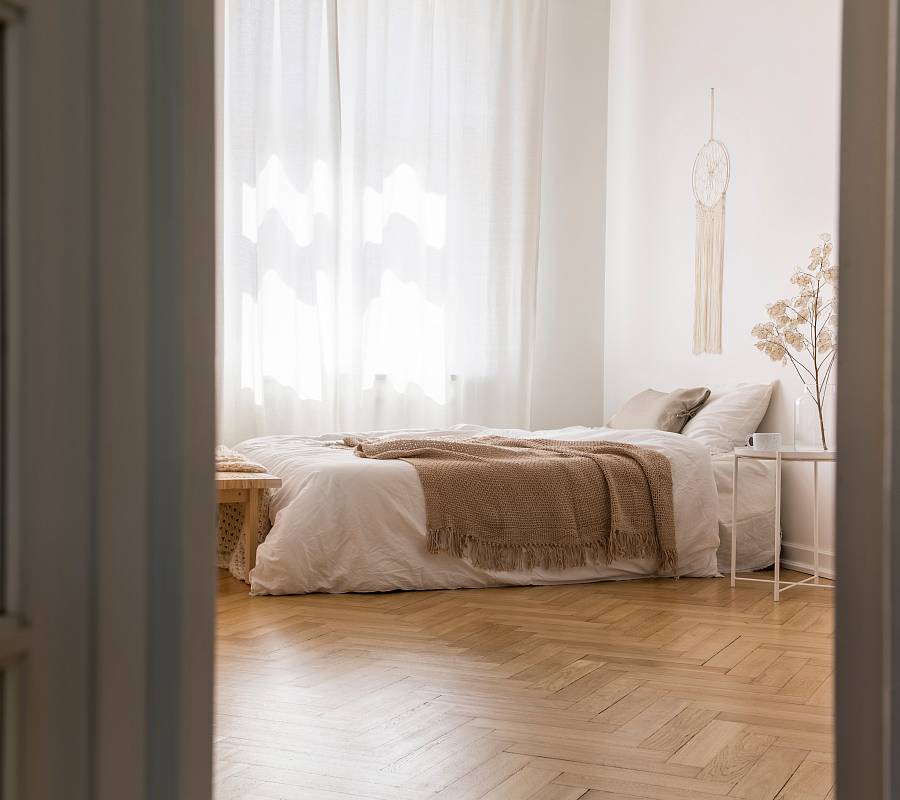 Blick in ein helles Schlafzimmer mit natürlichem Fischgrätparkett, bodennahem Bett, sanften Textilien und ruhiger Atmosphäre.