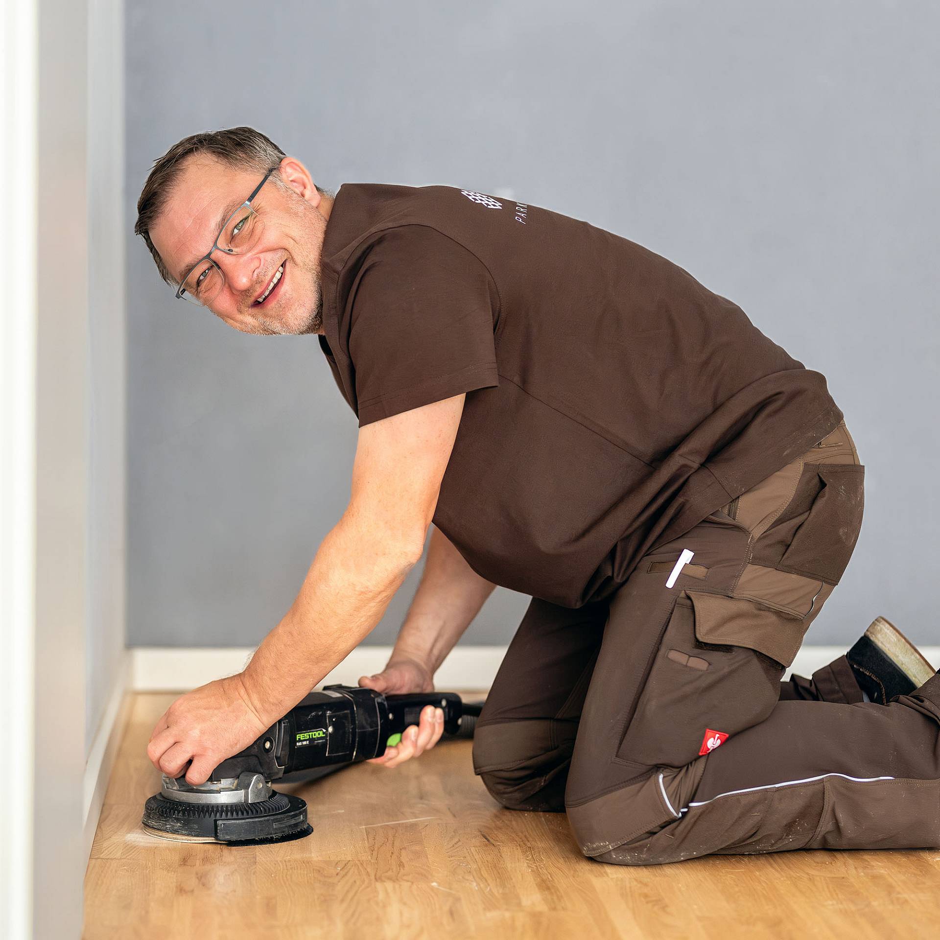 Parkettleger schleift Holzboden kniend mit Randschleifer im Innenraum