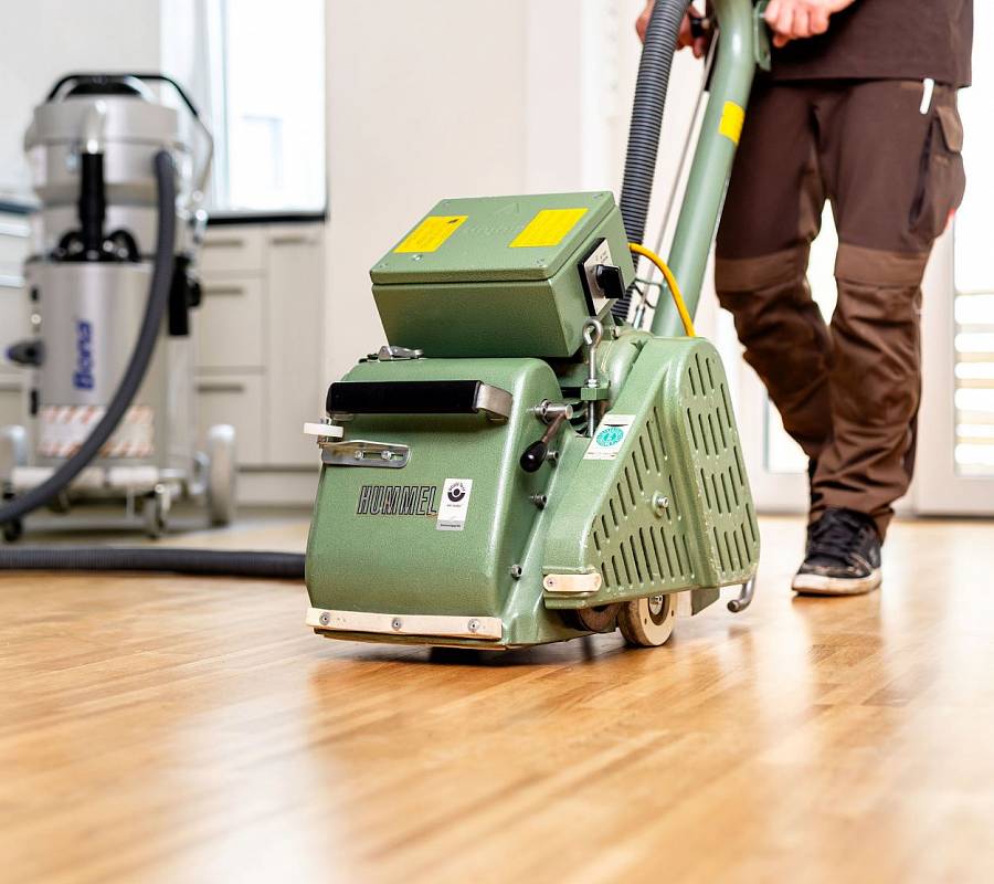 Mitarbeiter schleift Holzboden mit einer grünen Bodenschleifmaschine; im Hintergrund steht ein Industriesauger.