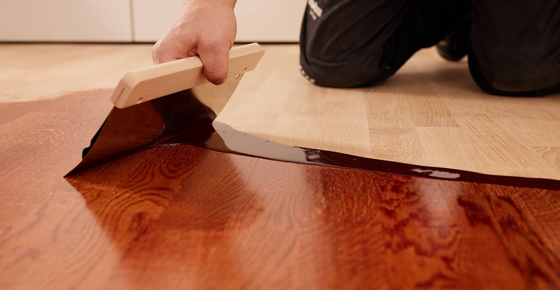 Gleichmäßig gefärbter Holzboden mit warmen Brauntönen, auf den Sonnenlicht weiches Licht wirft. Ein Handwerker verstreicht mit der Spachtel die Färbung.
