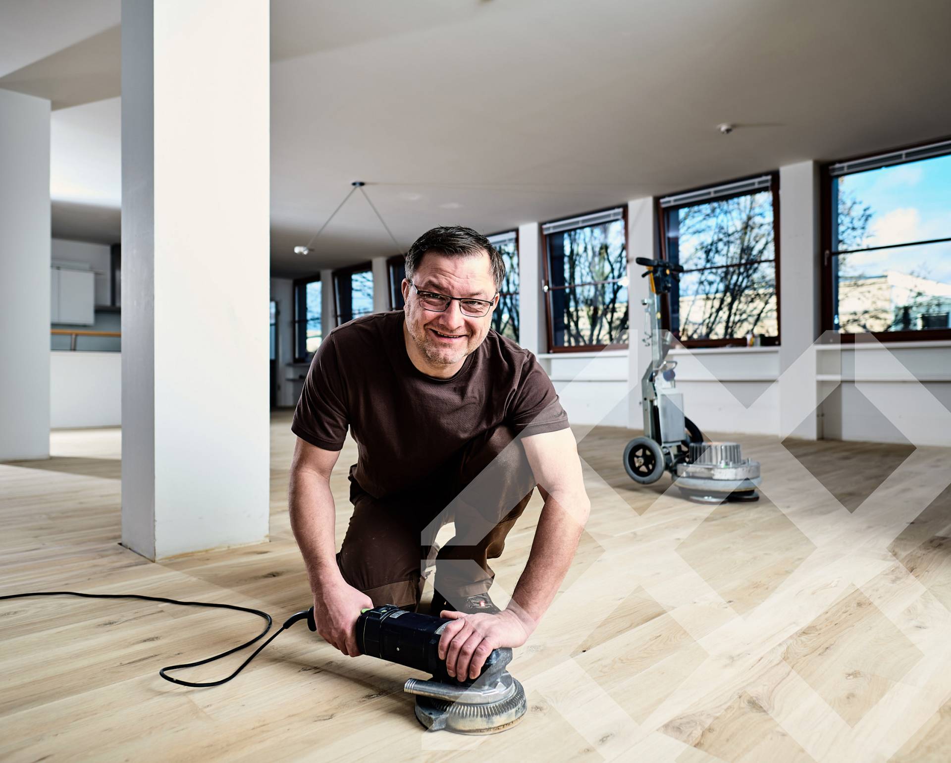 Parkettleger arbeitet kniend mit Schleifmaschine auf hellem Holzboden in großzügigem, lichtdurchflutetem Raum.
