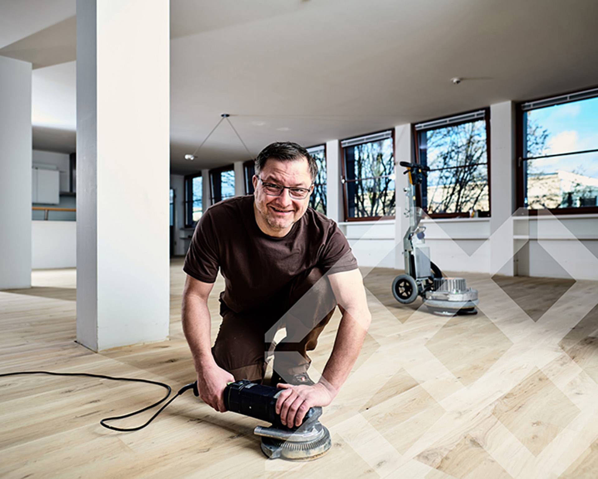 Parkettleger arbeitet kniend mit Schleifmaschine auf hellem Holzboden in großzügigem, lichtdurchflutetem Raum.