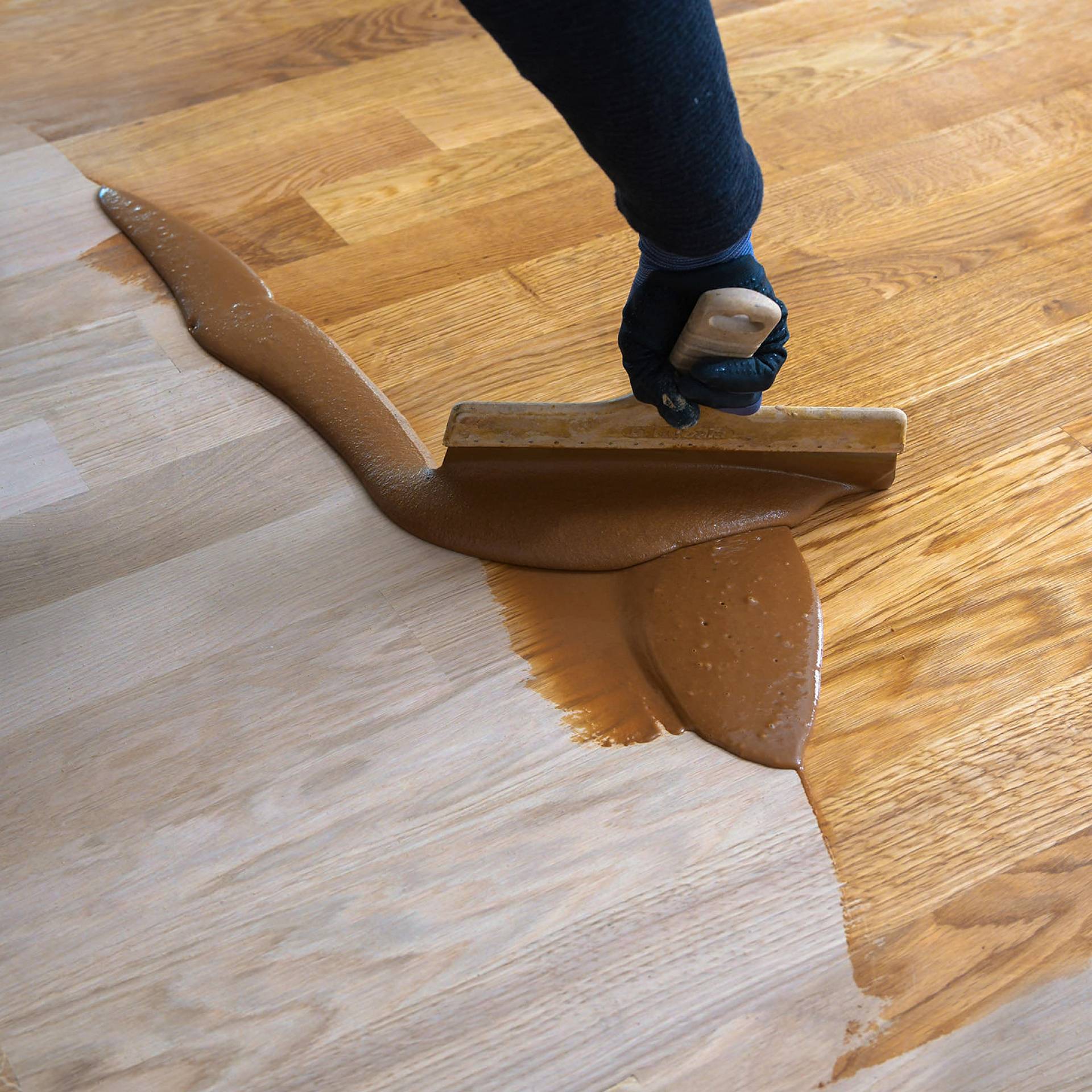 Hand mit Spachtel verteilt braune Beschichtungsmasse auf Parkettboden zur Oberflächenbehandlung.