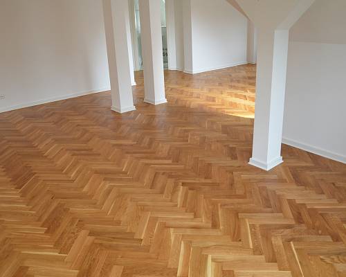 Heller Wohnbereich mit warmem Eichen-Fischgrätparkett und weißen Stützen.