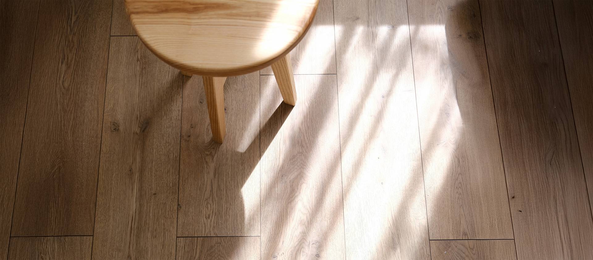 Sonnenlicht fällt durch ein Fenster auf einen hellen Holzboden; ein runder Holzstuhl steht teilweise im Licht.
