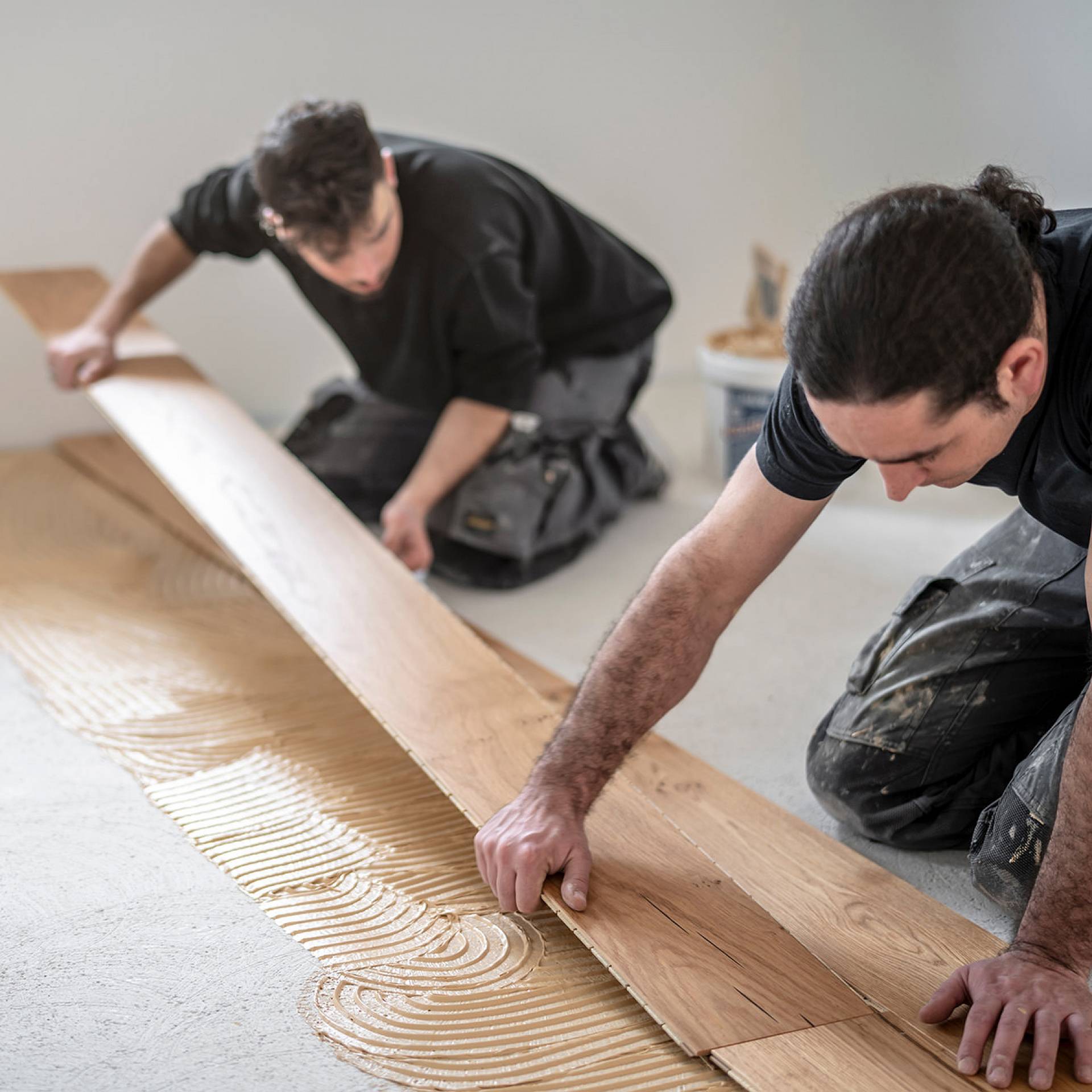 Zwei Handwerker verlegen sorgfältig Holzboden auf frisch aufgetragenem Kleber.