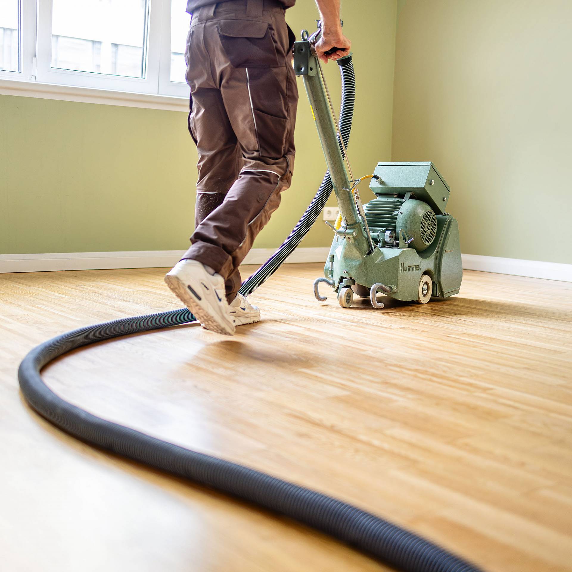 Ein Handwerker schleift einen Holzboden mit einer professionellen Parkettschleifmaschine und angeschlossenem Absaugschlauch.
