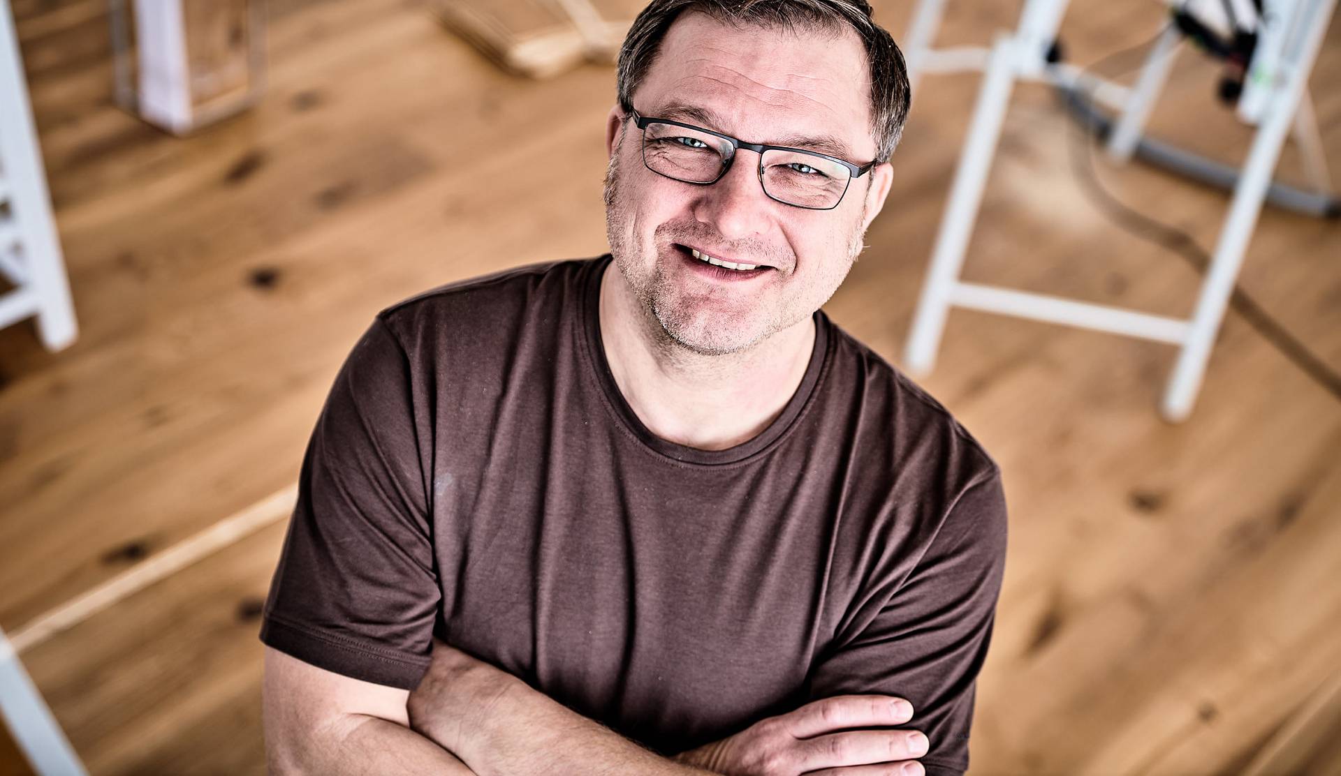 Lächelnder Mann mit Brille und verschränkten Armen steht auf einem Holzfußboden und blickt in die Kamera