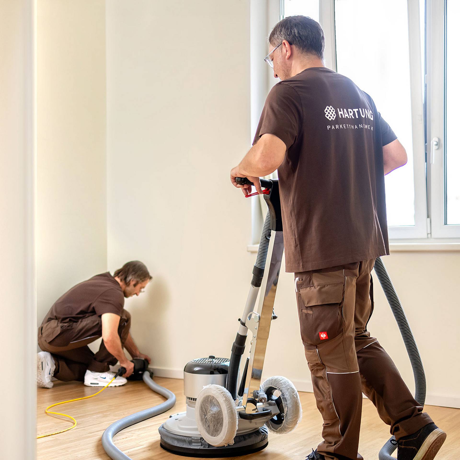 Zwei Handwerker schleifen und bearbeiten einen Holzboden mit Maschinen in einem Innenraum.