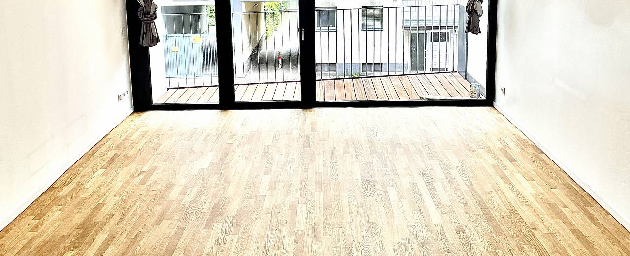 Leerer Raum mit großen Balkontüren und hellem, frisch geschliffenem Holzboden mit gleichmäßiger Oberfläche.