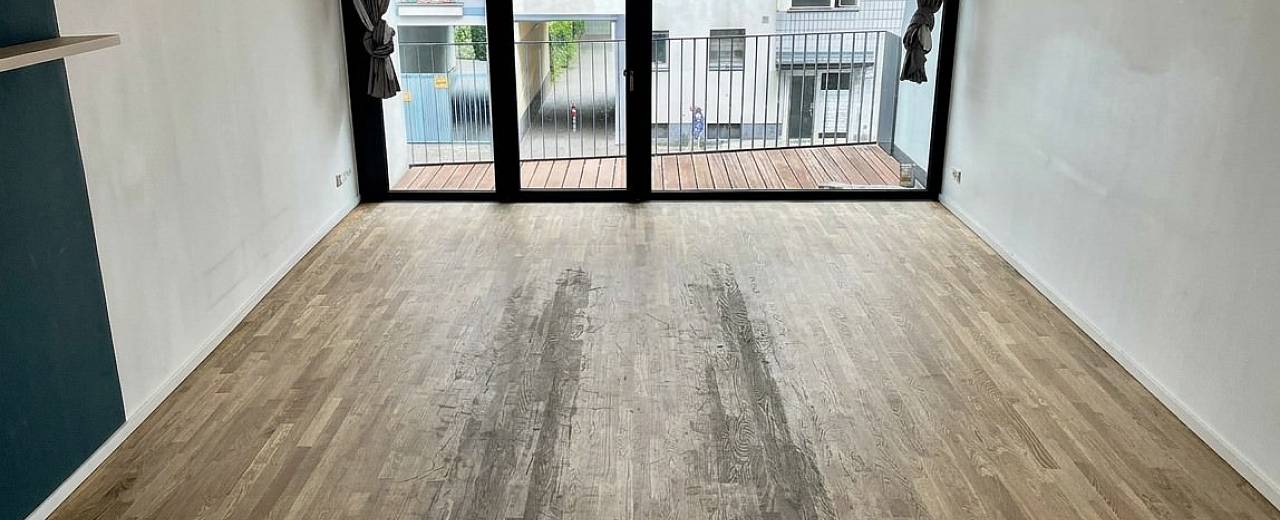 Leerer Raum mit großen Balkontüren und grauem, abgenutztem Holzboden mit sichtbaren Gebrauchsspuren.