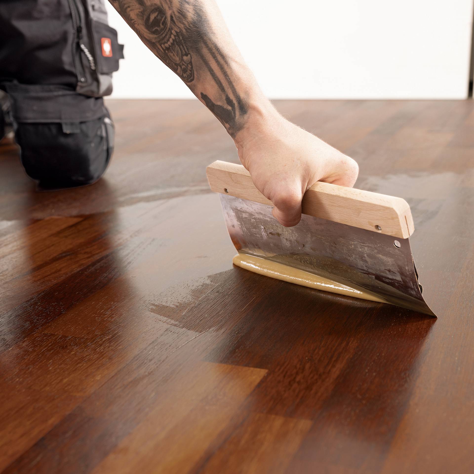 Hand trägt mit Spachtel Öl auf Holzparkett auf; daneben steht eine Dose Parkettöl.