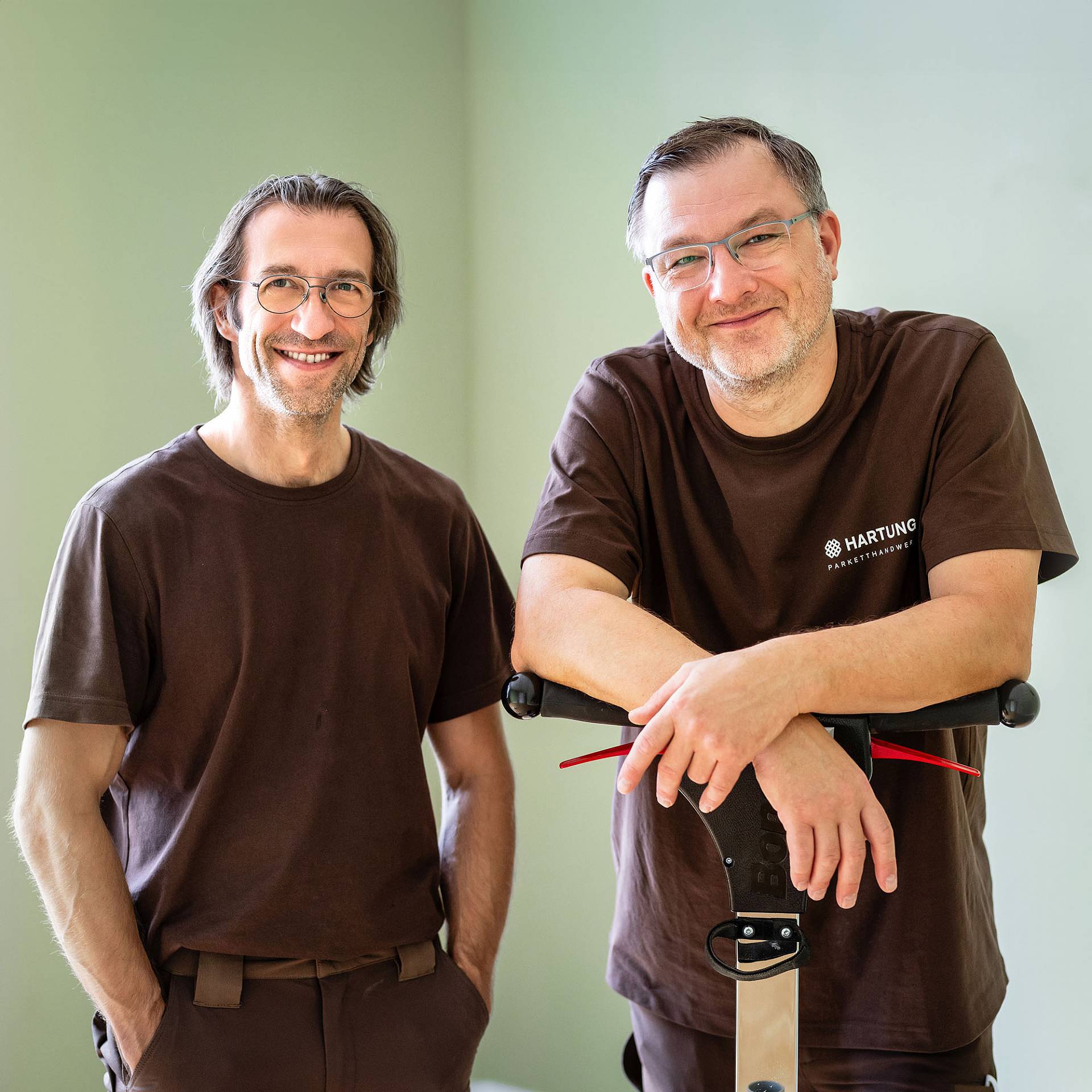 Zwei Männer in braunen Arbeits-T-Shirts stehen nebeneinander und lächeln; einer lehnt sich auf ein Werkzeug.