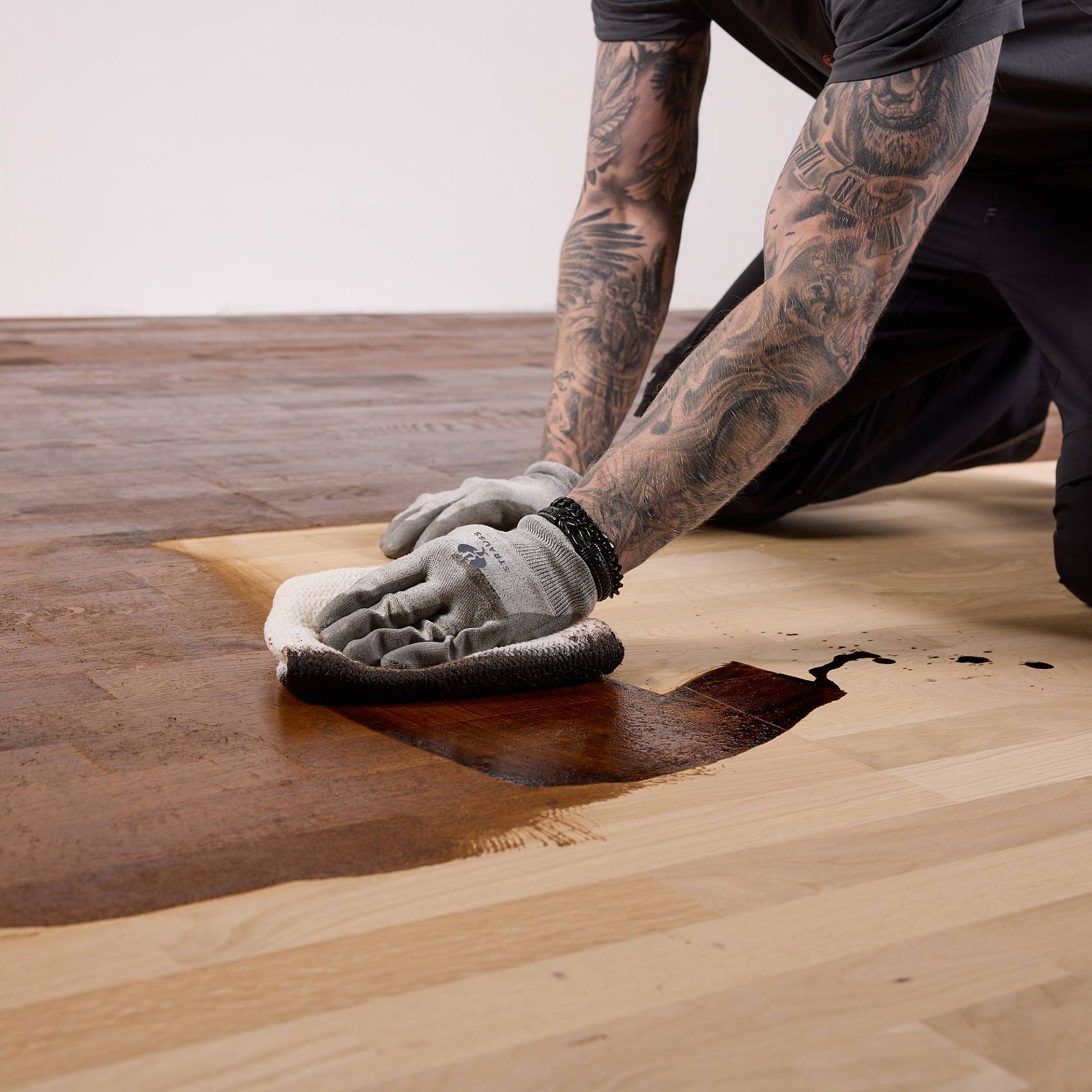 Handwerker mit tätowierten Armen und Arbeitshandschuhen arbeite dunkle Färbung mit einem Microfasertuch in den Holzboden ein.