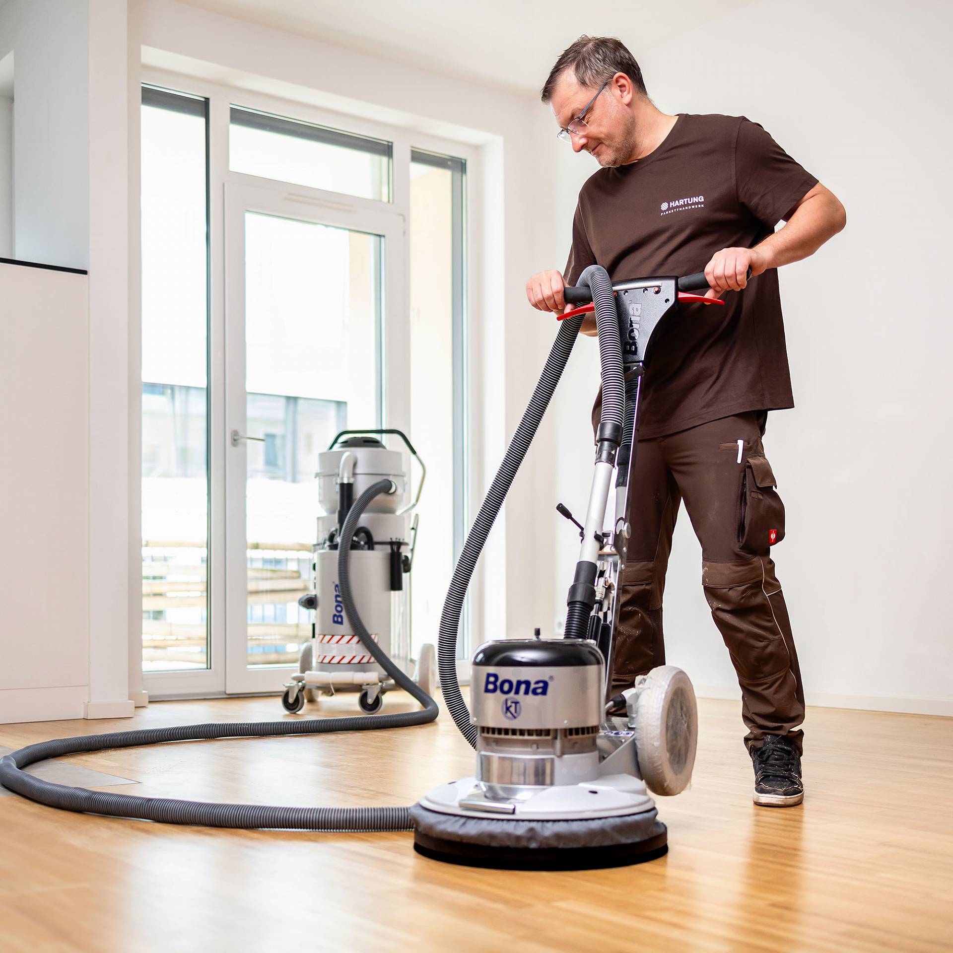 Handwerker bedient Poliermaschine auf hellem Holzboden vor Fensterfront in leerem Raum