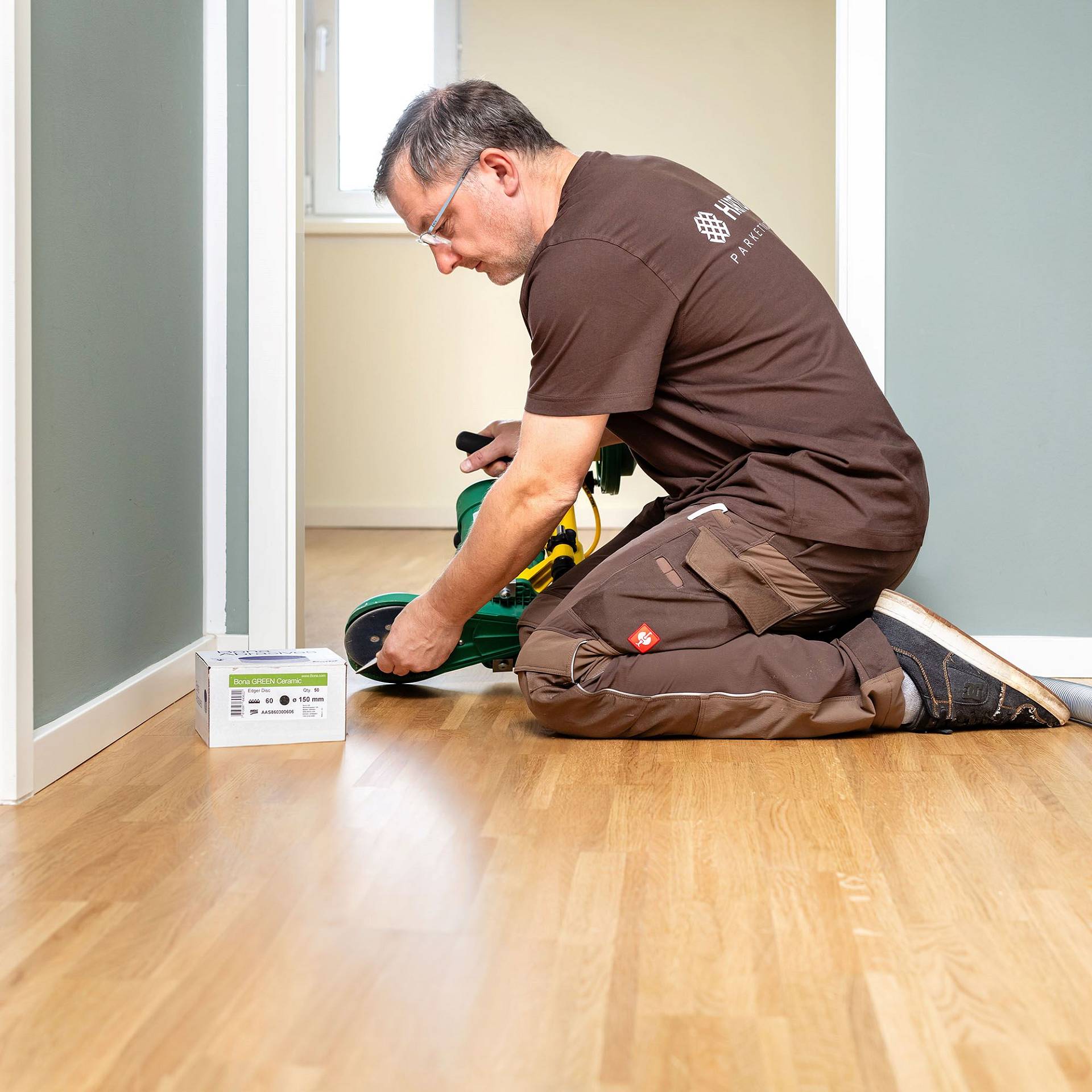 Handwerker kniet auf Holzboden und bedient ein Elektrowerkzeug beim Verlegen oder Bearbeiten von Dielen.