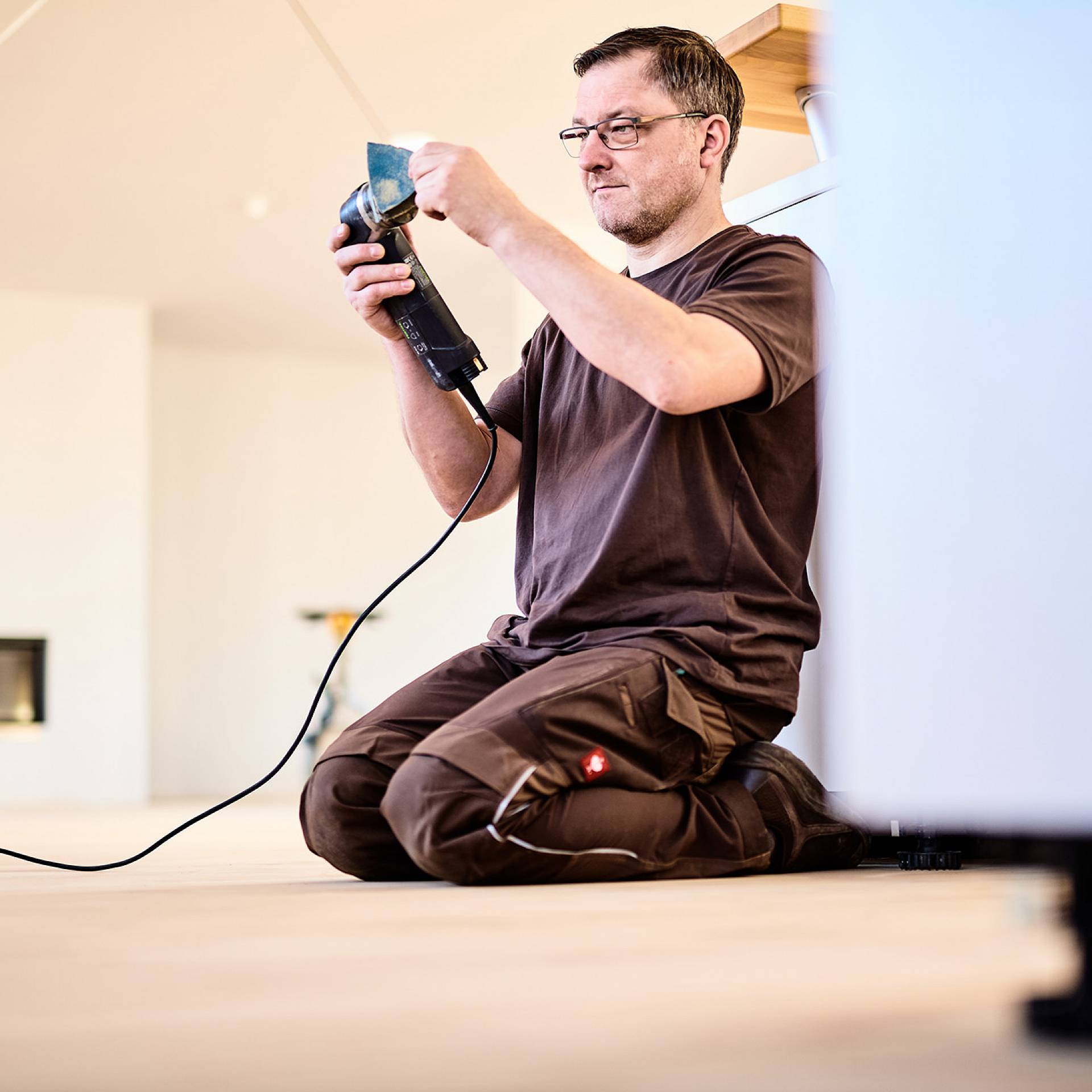 Handwerker kniet auf Holzboden und bedient ein Elektrowerkzeug beim Verlegen oder Bearbeiten von Dielen.