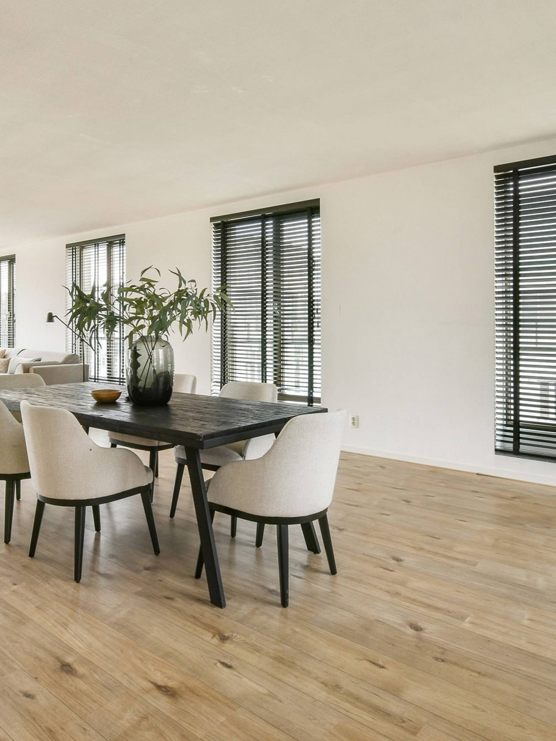 Modernes Esszimmer mit Holzdielenboden, dunklem Tisch, Polsterstühlen, Pflanzen und großen Fenstern mit Jalousien.