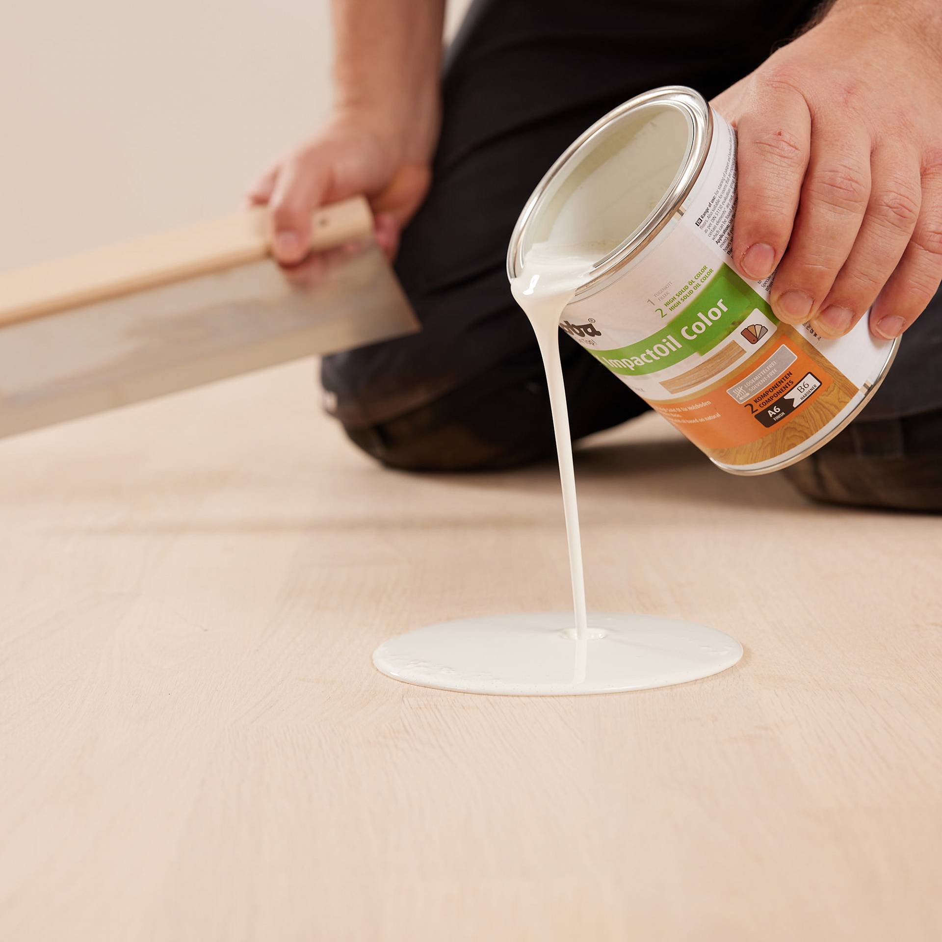 Handwerker gießt pigmentiertes Öl auf den Holzboden. In der anderen Hand hält er eine Spachtel, um es gleichmäßig zu verteilen.