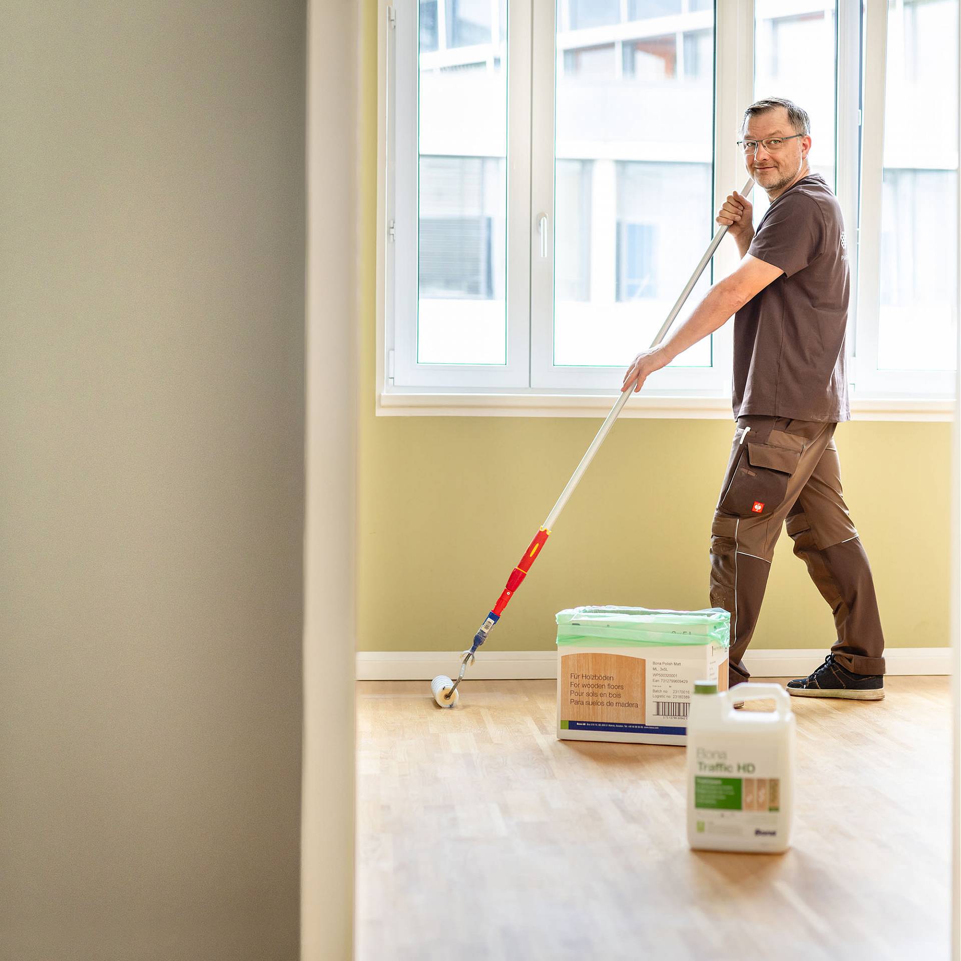 Handwerker trägt mit Farbrolle Schutzmittel auf hellen Holzboden in einem leeren, lichtdurchfluteten Raum auf.