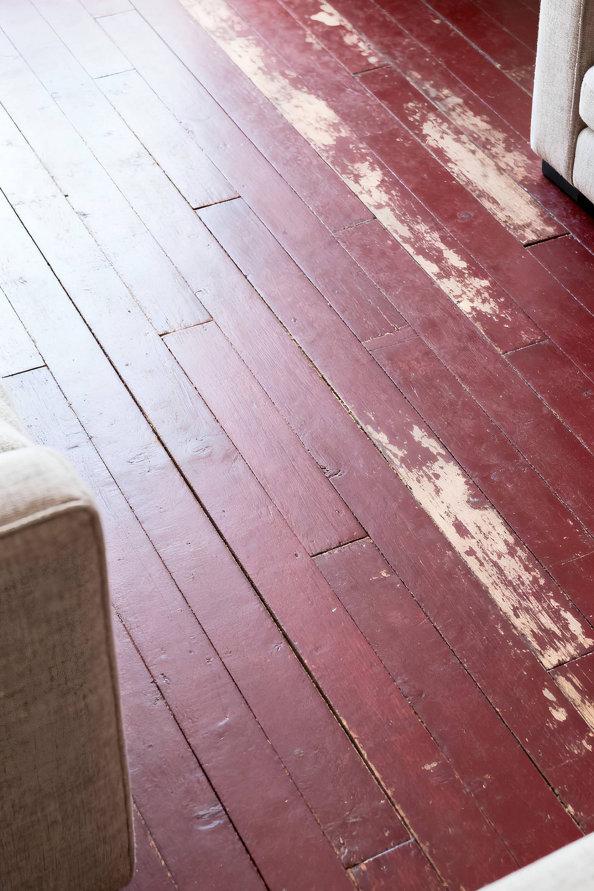 Abgenutzter Ochsenblut-Dielenboden mit abgeplatzter Farbe und sichtbaren Gebrauchsspuren, teilweise von Sofakanten gerahmt.