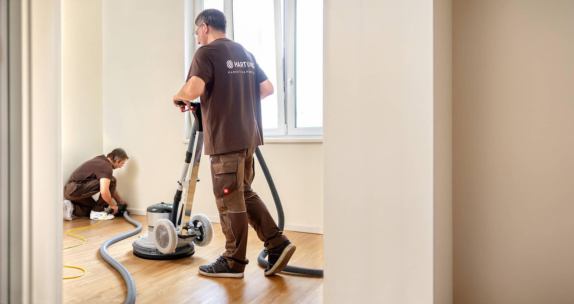 Zwei Handwerker schleifen mit einer Bodenschleifmaschine einen Holzboden in einem hellen Raum.
