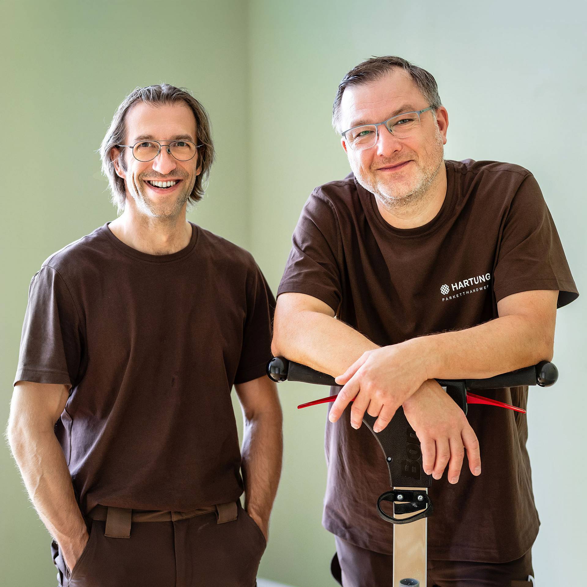 Zwei lächelnde Handwerker in braunen Shirts stehen in einem hellen Raum neben einer Schleifmaschine.
