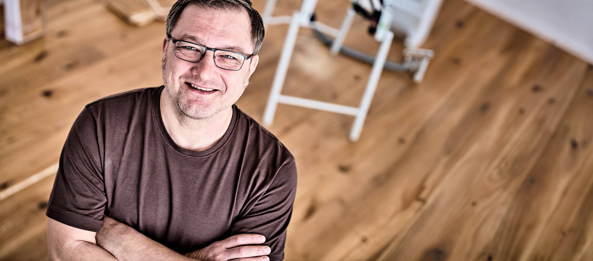 Lächelnder Mann mit Brille und verschränkten Armen auf Holzboden, Leiter im Hintergrund, Blick nach oben