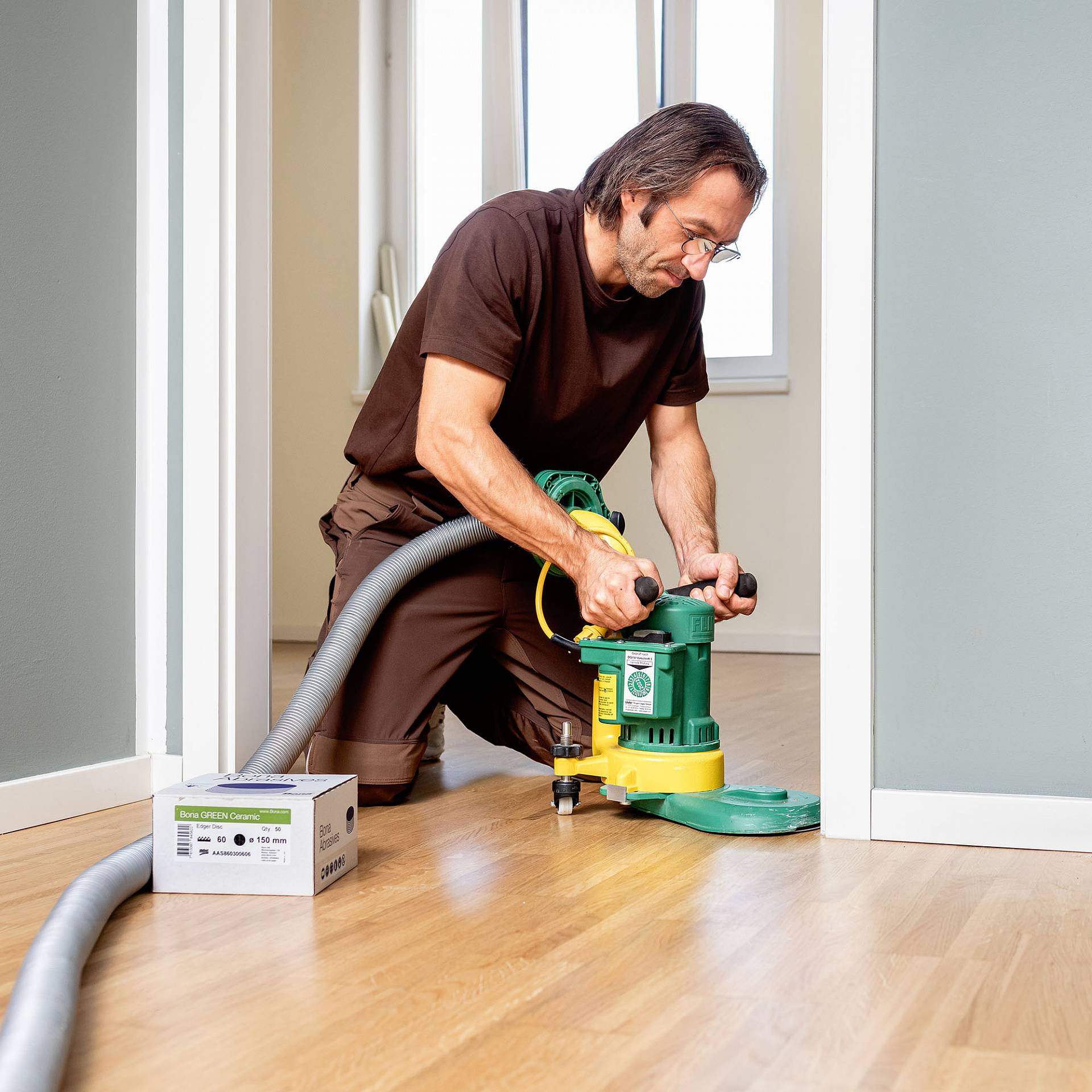 Parkettleger schleift Holzboden randnah mit Kanten­schleifmaschine im Innenraum